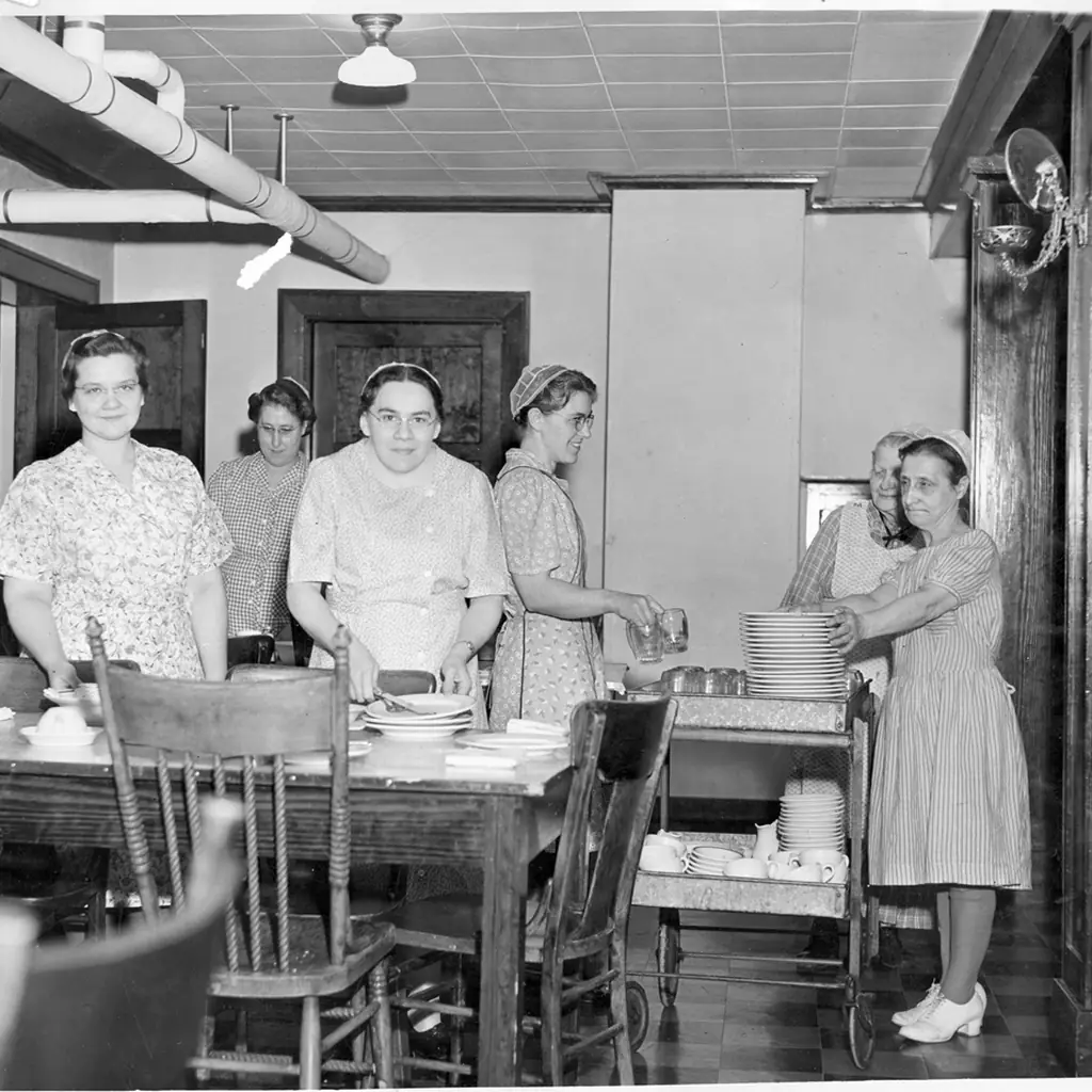 Women clearing dishes from a long kitchen table in 1951.